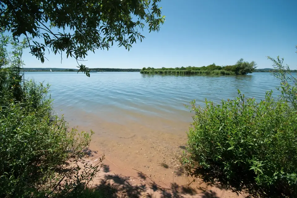Kleine Sandbucht zwischen Büschen, klares flaches Wasser und eine bewachsene Insel unter blauem Himmel
