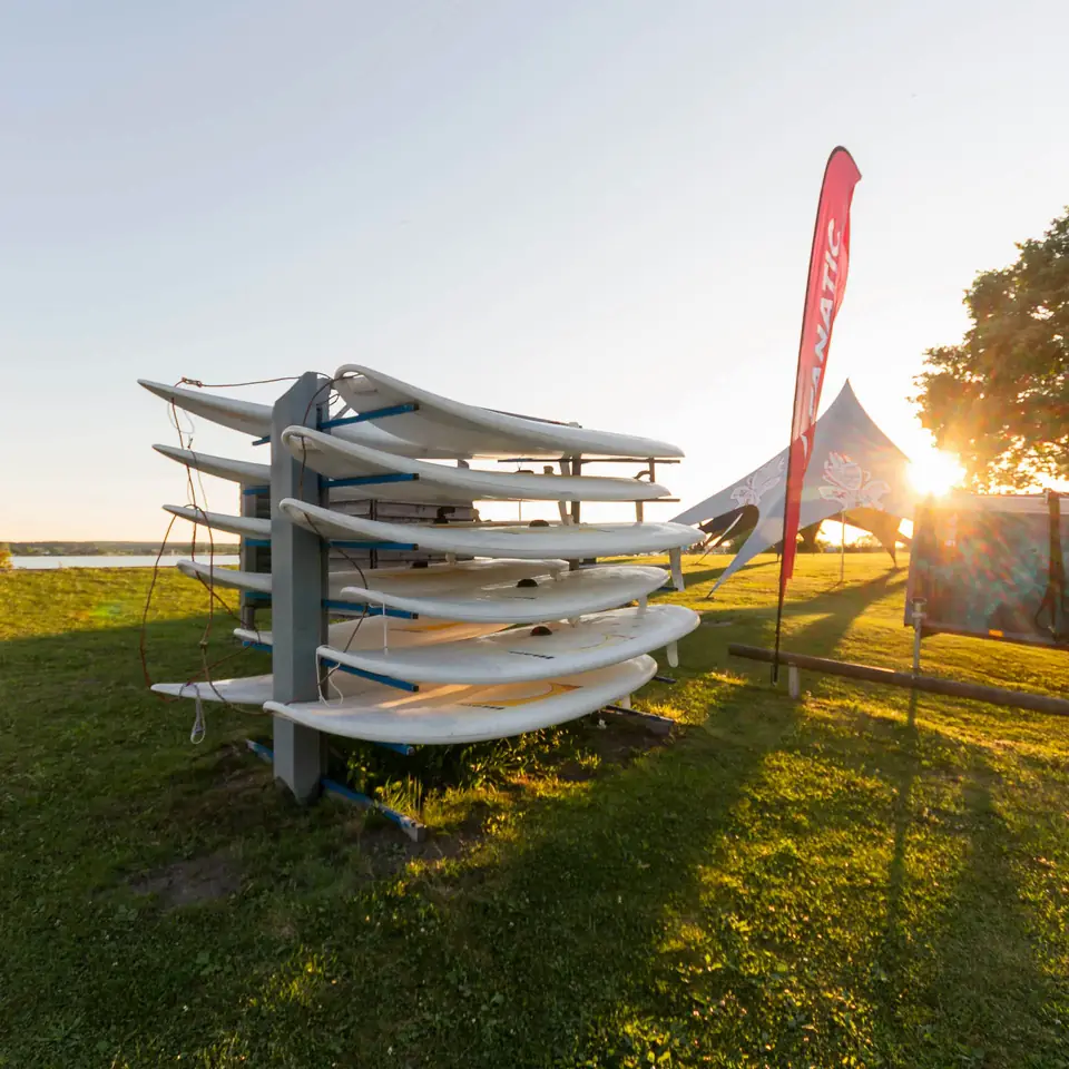 Gestell mit Surfbrettern auf einer Wiese am Ufer, dahinter Fahne und Zelt im tiefen Sonnenlicht