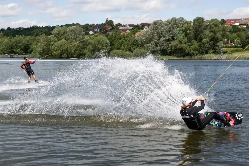 Zwei Wakeboarder an der Seilbahn ziehen Gischt über das Wasser, dahinter Bäume und Häuser am Ufer